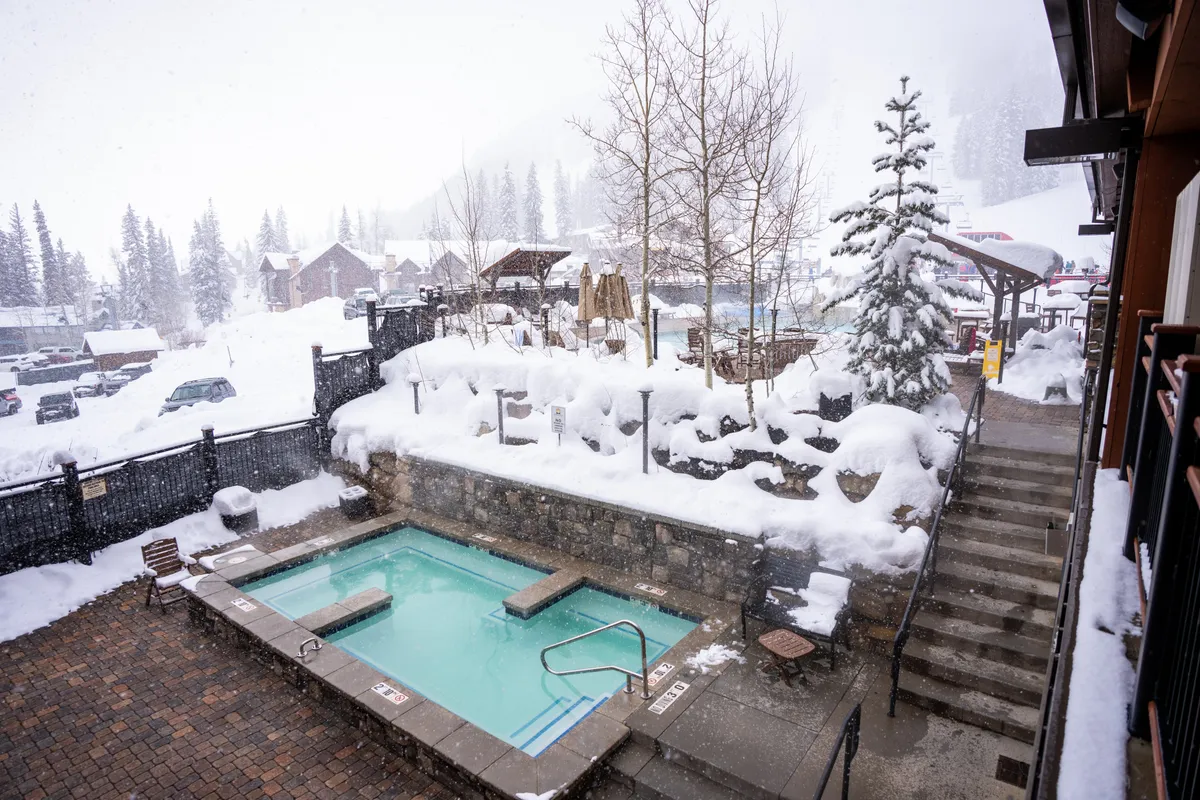 A tranquil snow-covered scene with a hot tub nestled among trees and steps leading down, offering a peaceful space to unwind after an active day on the mountain. The view extends to the surrounding winter landscape, inviting relaxation.