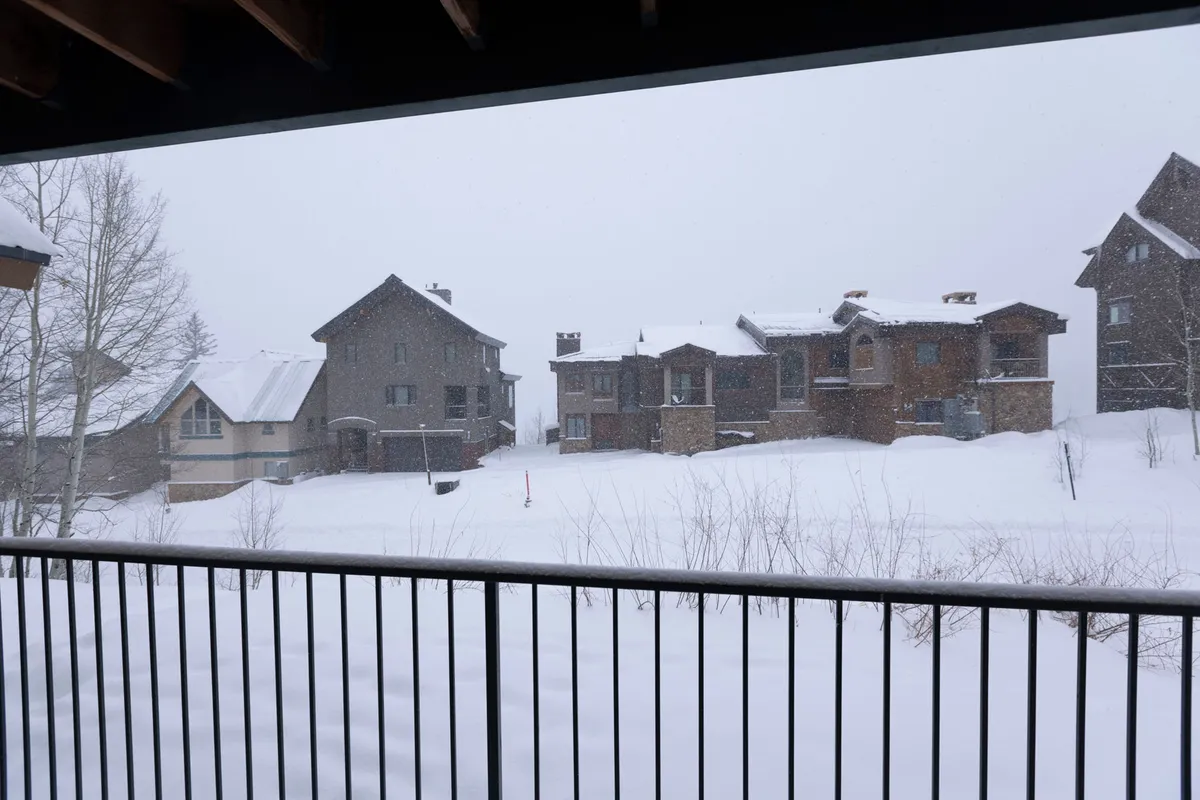 A peaceful wintertime view featuring a snow-covered landscape with houses sporting snow-laden roofs and tree branches dusted in white. The mood is tranquil and cozy, capturing the calm of watching snowfall from inside, possibly while enjoying a warm drink after a day on the slopes.