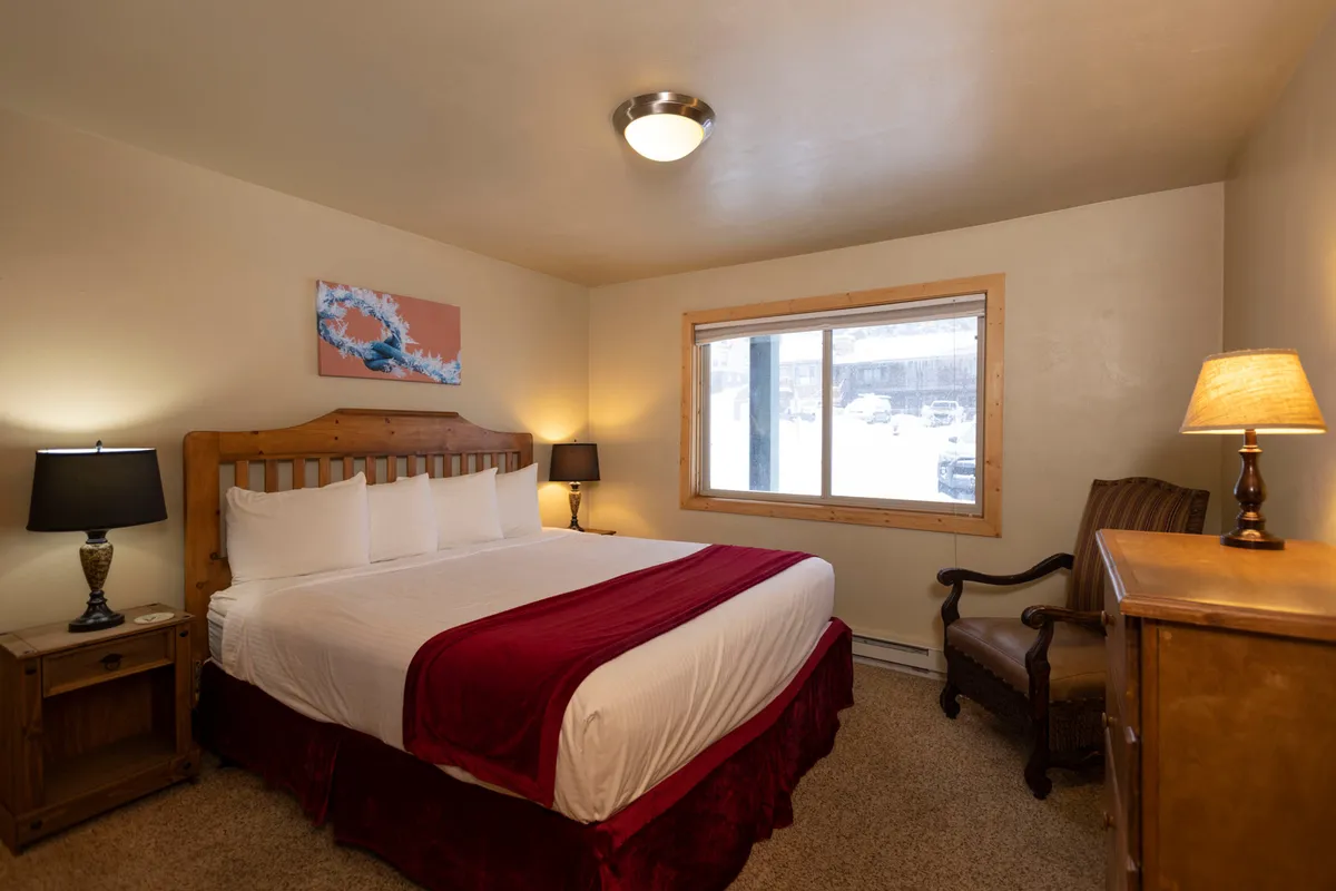 A warmly lit interior featuring a bed positioned near windows that let in plentiful natural light. Artwork and a cozy lamp contribute to an inviting mood, and the windows provide glimpses of the mountain scenery outside.