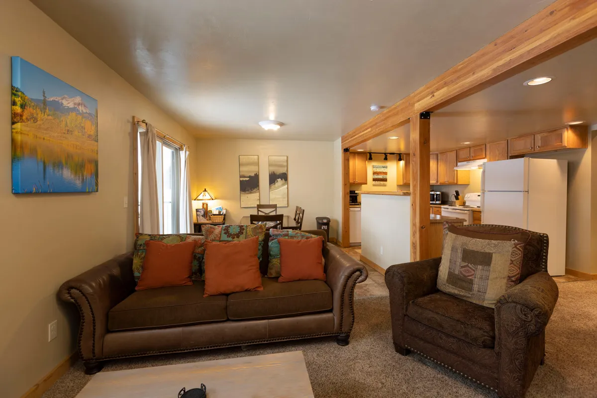 The image showcases a warm, inviting interior with a plush sofa featuring colorful pillows, a modern dining table set for gatherings, a fully equipped kitchen in the background, and vibrant artwork on the wall. The space exudes comfort and style, ideal for relaxing, socializing, and enjoying home-cooked meals after a day in the mountains.