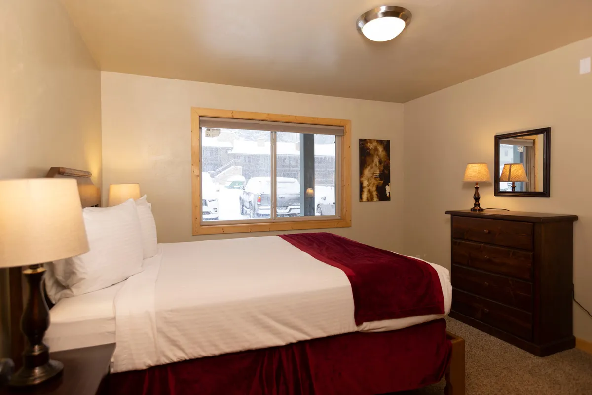 The image showcases a bedroom with white linens accented by a red throw, a window framing a snow-covered vista, and a dresser with a mirror. A lamp on the nightstand adds to the cozy, peaceful mood, while the uncluttered decor highlights the restful character of the space.