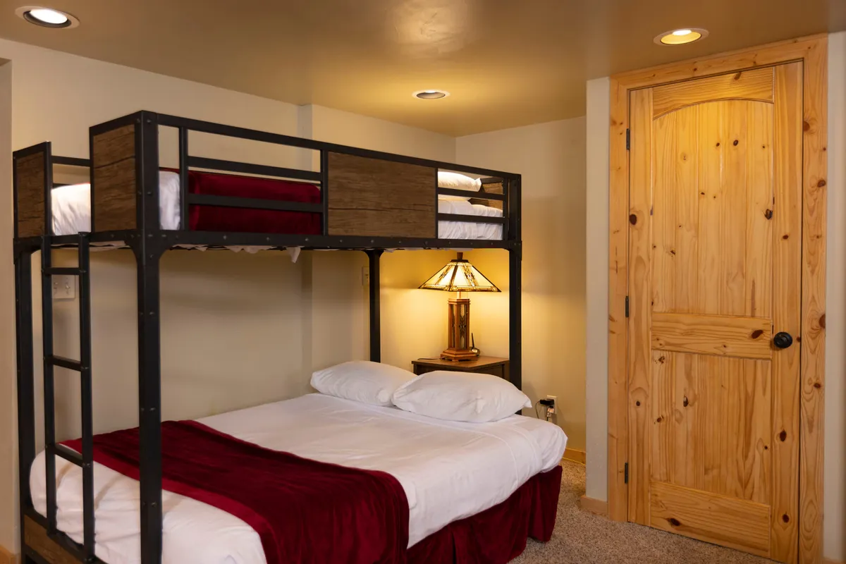 A family-friendly bedroom featuring a double bed and a bunk bed, accompanied by a table lamp and a wooden door. The room is designed for comfort, accommodating families or small groups, and offers a laid-back, intimate setting for restful sleep and quiet activities like reading.