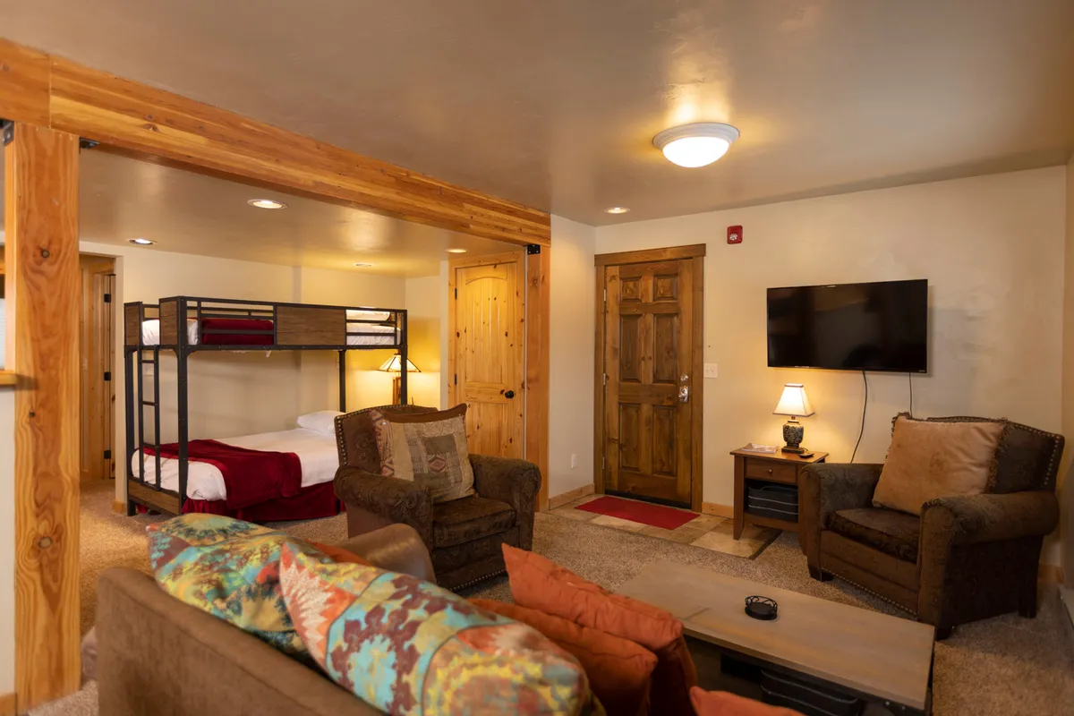 This interior image features a living space with a bunk bed, seating area, television, and entrance door. The bunk bed includes a queen futon on the bottom and a twin on top, while the seating area and TV create a cozy spot for relaxing and socializing. The lighting and arrangement suggest a welcoming area perfect for group gatherings or winding down after outdoor adventures.