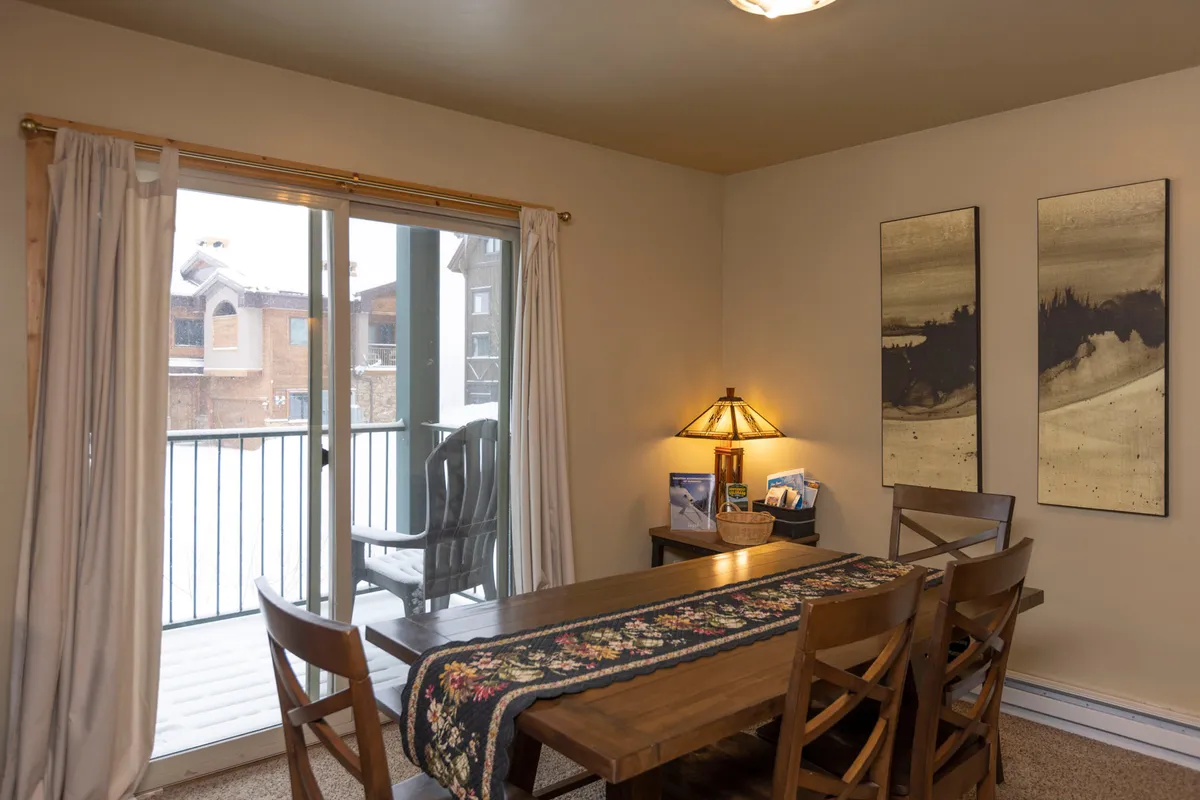 A view of a warmly lit dining area featuring a large table set near a sliding glass door, with artistic decor on the wall and a lamp adding to the inviting ambiance. The space suggests room for group gatherings and easy access to the outdoors, hinting at the larger alpine environment beyond.