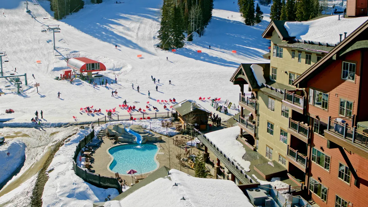 Overlooking the resort exterior, this image features the snowy ski slopes, ski lift, swimming pool, and hotel against a picturesque mountain backdrop. The scene bustles with winter activity and hints at both daytime adventure and après-ski relaxation.