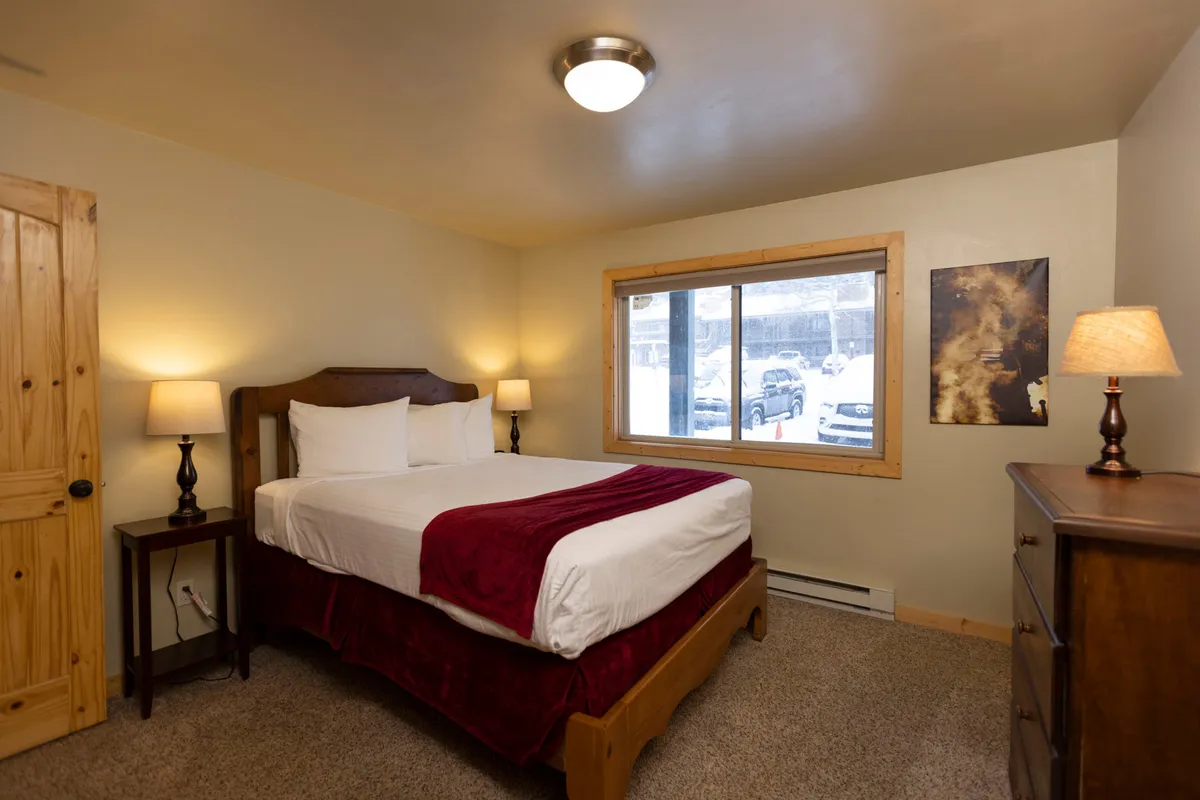 This image features a cozy bedroom with a bed adorned with a red throw, positioned near a large window. Nightstands with warm lamps flank the bed, while natural light fills the space, creating a warm and restful ambiance. The inviting decor emphasizes both comfort and privacy, making it an ideal setting for relaxation after a busy day.